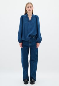 Kék, texturált blúz V-nyakkal és fodros szélekkel, párosítva magas derekú, laza szárú sötét farmernadrággal. A lábbelik fekete talppal rendelkeznek.