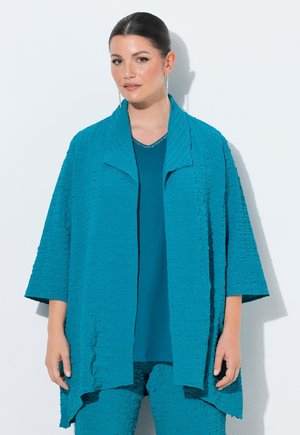 Vrouw draagt een getextureerd teal jasje, bijpassende broek en top, staat tegen een witte achtergrond, met lange oorbellen en glad haar.