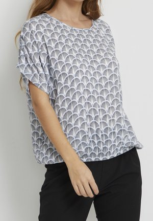 Femme portant un chemisier blanc ample avec un motif noir en forme d'éventail et un pantalon noir, debout devant un fond clair uni.