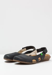 Chaussures en cuir noir avec un design à découpes, une sangle réglable et une semelle texturée marron. Accent vert notable sur le côté.