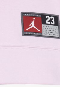 Tessuto rosa chiaro con un'etichetta nera e rossa che recita "BRAND OF FLIGHT 23" con un logo bianco in silhouette e dettagli testuali.