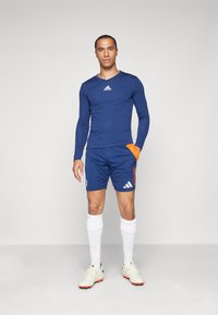 Maglietta sportiva a maniche lunghe di colore navy con logo bianco, abbinata a pantaloni corti blu con accenti arancioni e calzettoni bianchi al ginocchio. Scarpe da calcio di colore chiaro.