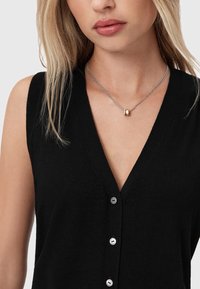 Kobieta w czarnej bezrękawnikowej bluzce z dekoltem w serek, z guzikami oraz srebrnym naszyjnikiem z dwoma małymi zawieszkami.