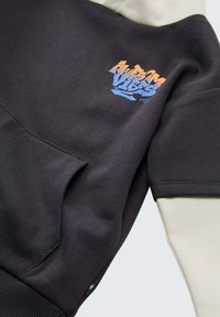 Felpa grigia scuro con maniche crema, caratterizzata da un design grafico con testo arancione e blu. Comprende una tasca frontale e polsini a coste.