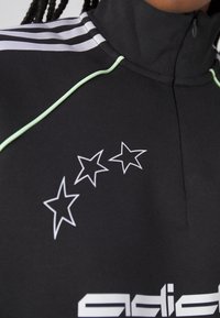 Svart högkragad sweatshirt med gröna detaljer, tre vita stjärnor på bröstet och vit logotyp längst ner; har sportigt, texturerat tyg.