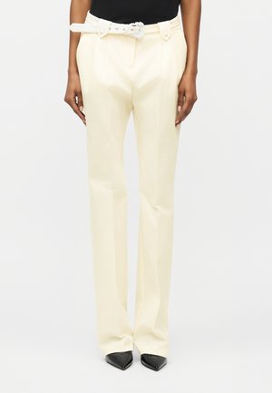 Persona que lleva pantalones de talle alto color crema, con pliegues, un cinturón blanco con hebilla decorativa y zapatos negros puntiagudos.