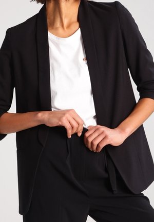 Person wearing black blazer and pants with white t-shirt, adjusting or fastening the waistband of the pants.