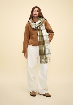 Giacca in camoscio marrone, pantaloni bianchi e una sciarpa fantasia con strisce verdi, marroni e beige, con frange alle estremità.