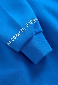 Blauwe sweatshirt met ribbelmanchetten. Voorzien van witte tekst met coördinaten: "51.5072° N, 0.1276°". Zachte, gestructureerde stof.