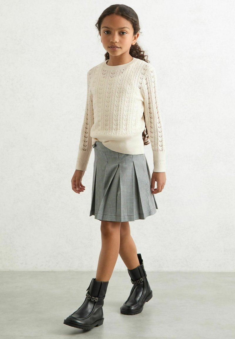 Strick- cremfarbener Pullover mit Durchbruchsdesign, grau karierter plissierter Rock und schwarze Lederstiefeletten mit Kettendetails.