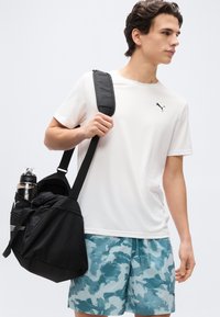 Νεαρός άνδρας που φοράει λευκό μπλουζάκι Puma και μπλε σορτς με σχέδια, κρατώντας μια μαύρη τσάντα γυμναστηρίου με ένα μπουκάλι νερό στην πλαϊνή τσέπη.