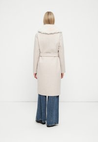 Cămașă bej deschis cu un guler din blană sintetică, legată în talie, cu un croi drept, ajungând până la mijlocul coapselor, asortată cu pantaloni din denim evazați.