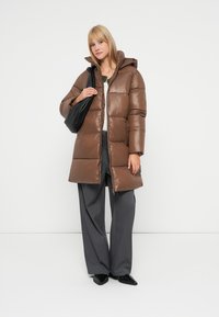 Blonde vrouw in een bruine gewatteerde jas, grijze wijde broek, zwarte puntige schoenen en met een zwarte schoudertas tegen een witte achtergrond.