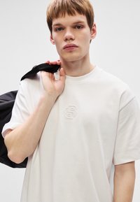 Ανοιχτό χρώματος μπεζ μπλούζα με στρογγυλή λαιμόκοψη και ανάγλυφο λογότυπο στο μπροστινό μέρος. Το ύφασμα φαίνεται λείο, φορεμένο με μια μαύρη θήκη πάνω από τον ώμο.