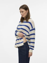 Pulover tricotat cu dungi în crem cu nuanțe de albastru, având o croială lejeră, mâneci largi și material texturat. Combinat cu pantaloni din denim închis la culoare.