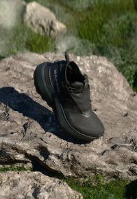 Schwarzer Wanderschuh mit strukturiertem Obermaterial, integriertem Schnürsystem und dicker Sohle, auf einer felsigen Oberfläche mit grüner Umgebung im Hintergrund.