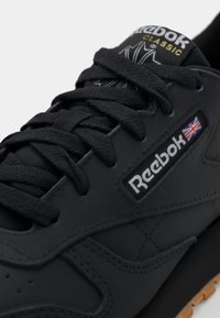 Svarte Reebok-sneakers har glatt skinn, tykke snører, en logo-lapp og en gummisåle, med diskret søm og minimalistisk design.