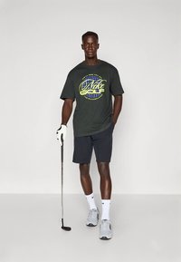 Μπλούζα κοντομάνικη σκούρου πράσινου χρώματος με κίτρινο και μπλε γράφημα "Nike Golf", συνδυασμένη με σκούρα μπλε σορτς, λευκές κάλτσες και γκρι αθλητικά παπούτσια. Κρατάει ένα γκολφ κλαμπ.