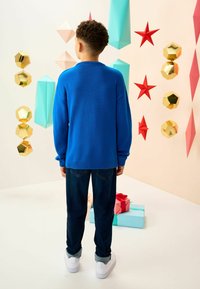 Pulover tricotat albastru cu mâneci lungi, asociat cu blugi închisi la culoare și adidași albi. Fundalul este decorat cu geometrice colorate și cutii de cadouri.