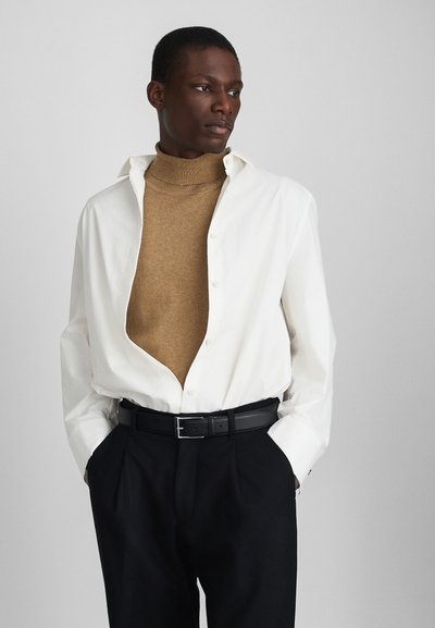 Camisa oversized branca sobre um suéter de gola alta castanho, combinada com calças escuras e um cinto preto. A camisa tem uma frente com botões.