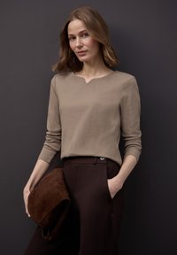 Beżowa prążkowana bluzka z długim rękawem i dekoltem w serek, zestawiona z ciemnobrązowymi spodniami. Modelka trzyma brązową kopertówkę zamszową. Tło w kolorze ciemnoszarym.