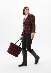 Donna in un blazer scozzese con colletto in stampa leopardata, che porta una borsa a tracolla nera e rossa, indossando pantaloni neri e stivali fino al ginocchio, cammina con sicurezza.