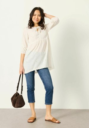 Junge Frau trägt eine weiße bestickte Tunika, blaue Jeans in Cropped-Länge, hellbraune Slide-Sandalen, hält eine braune Handtasche und lächelt mit der Hand am Kopf.