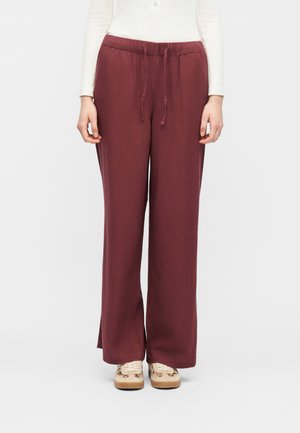 Donna che indossa pantaloni bordeaux larghi con coulisse e sneakers beige, mostrata dalla vita ai piedi su sfondo bianco.