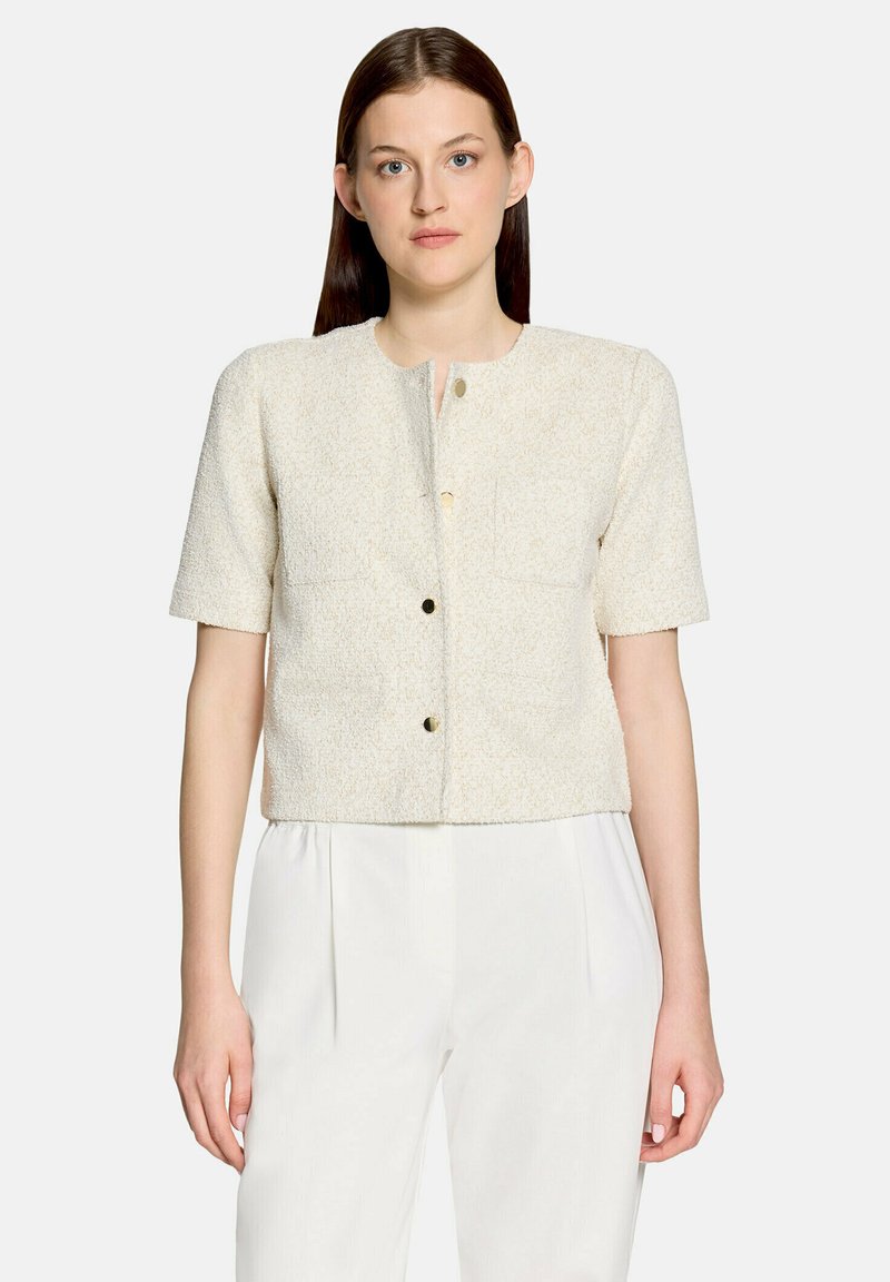 Femme portant une veste texturée crème à manches courtes avec des poches avant et des boutons sur un pantalon blanc taille haute, sur un fond uni.