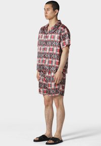 Camicia a maniche corte con colletto e pantaloni corti abbinati in un motivo grafico rosso e nero, realizzati in tessuto liscio. Indossati sandali neri a ciabatta.
