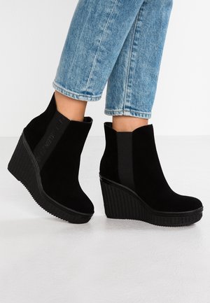 Sorte ankelstøvler med kilehæl i ruskind, båret med op rullede blå jeans på en hvid gulvbaggrund.