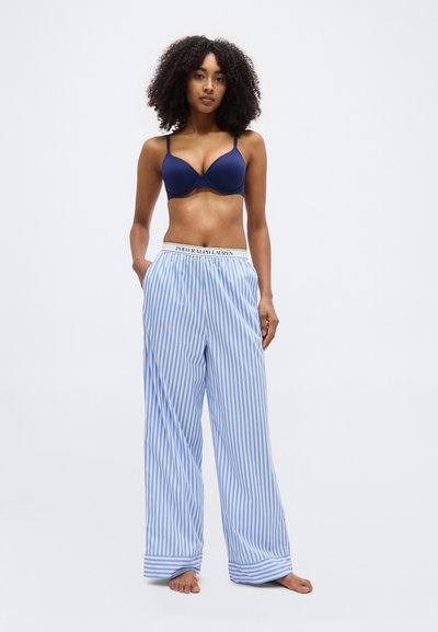 Femme aux cheveux bouclés portant un soutien-gorge bleu marine et un pantalon à larges jambes rayé bleu et blanc avec une ceinture "Polo Ralph Lauren", pieds nus, debout.