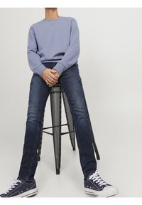 Sudadera azul clara con escote redondo, combinada con jeans azul oscuro. Sentado en un taburete de metal, lleva zapatillas de deporte marinas con cordones blancos.