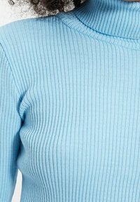 Jasnoniebieski sweter z prążkowanym wzorem i wysokim dekoltem; materiał wydaje się miękki i dopasowany, podkreślając kontury ciała.
