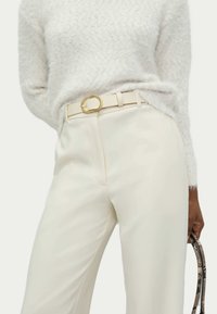 Un pantalon sur mesure couleur crème, avec une texture lisse, présentant une large ceinture avec une boucle circulaire en or et un doux pull gris clair.