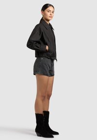 Kvinde står i sideprofil iført en sort jakke, mørke denimshorts og sorte støvler til midt på læggen mod en lys grå baggrund.