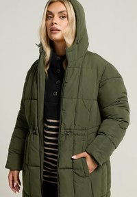 Olivgrön vadderad jacka med huva, som har dragkedja, snoddar och kontrasterande randiga innerlager. Texturerad kviltad design.