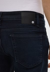 Jeans in denim blu scuro con una finitura testurizzata. Presentano un'etichetta in pelle nera sulla vita e dettagli nella tasca posteriore.