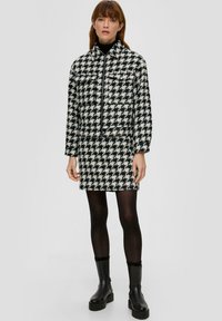 Mustriga houndstooth jakk ja seelik musta ja valge värviga, valmistatud tekstureeritud kangast, lühema lõike ja pikkade varrukatega. Mustad pahkluu saapad.
