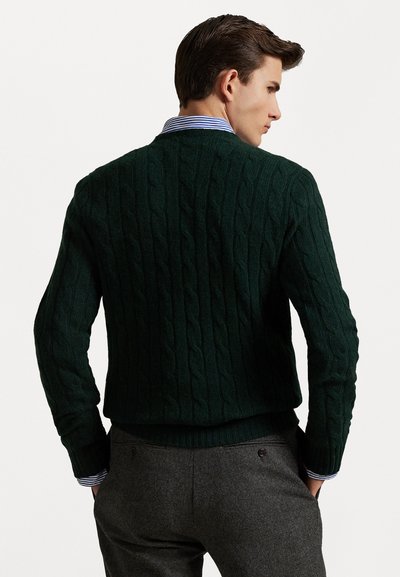 Pull en laine à mailles torsadées vert foncé avec un ourlet et des poignets côtelés, porté sur une chemise à col rayée bleu et blanc.