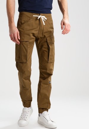 Man draagt bruine cargobroek met zijzakken en witte sneakers, staand tegen een effen lichte achtergrond.