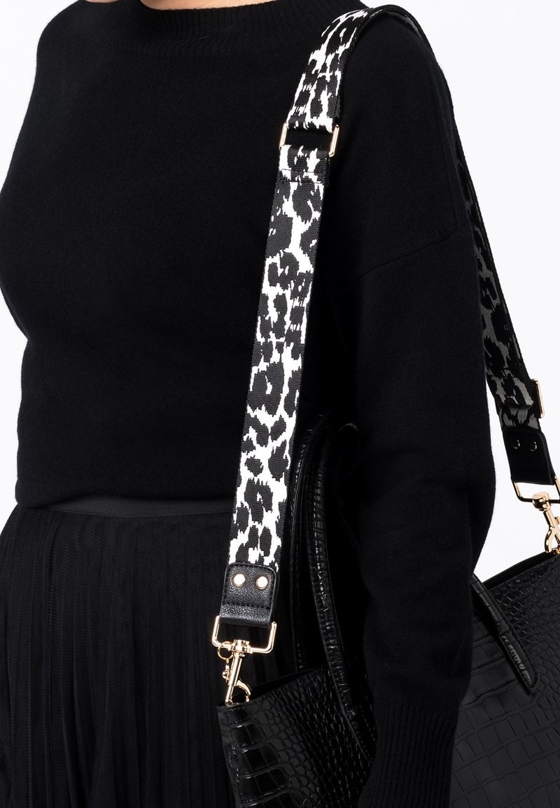 Personne portant un pull noir et une jupe, portant un sac à main noir texturé avec une bandoulière à imprimé animal noir et blanc.