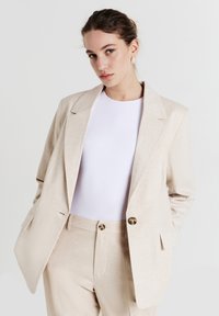 Woman wearing beige suit with oversized blazer and matching pants over white top, posing against plain light background.