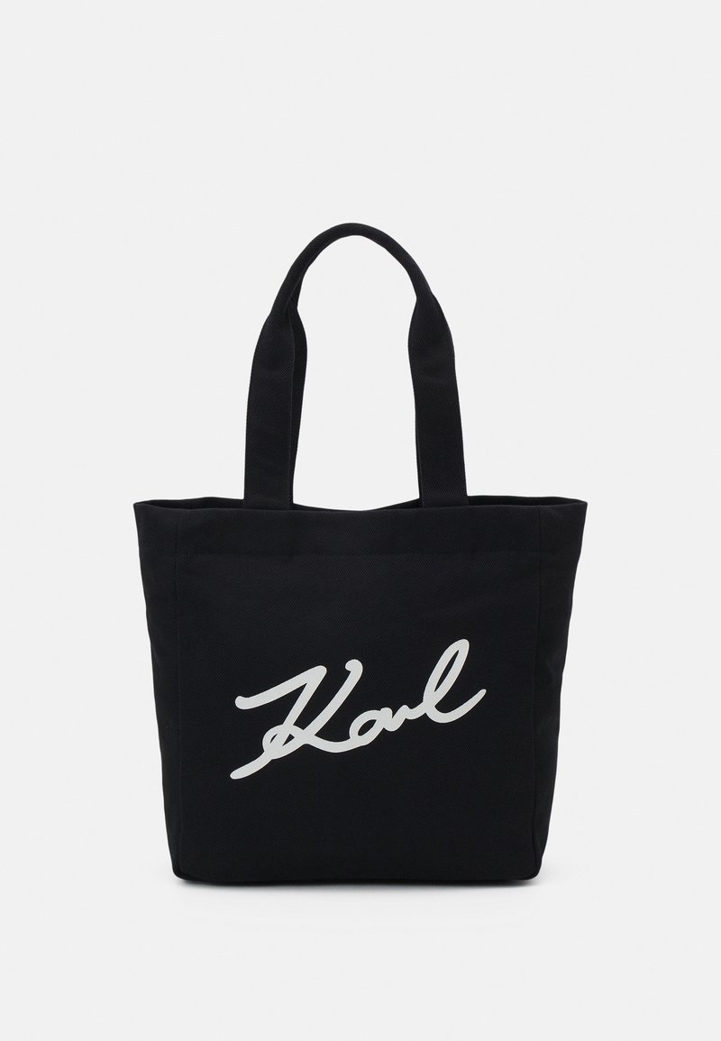 Sac cabas noir avec deux poignées et texte cursif blanc "Karl" sur le devant, présenté sur un fond uni gris clair.