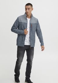 Fleece striukė su mėlynos ir pilkos dviejų tonų dizainu, užtrauktuku, aukštu apykakle ir kontrastinga užtrauktuko detale. Sukombinuota su tamsiomis džinsomis ir juodais batais.