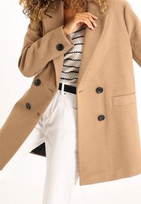 Mujer vistiendo un abrigo de doble botonadura en color beige, pantalones blancos y una blusa blanca de canalé con rayas onduladas negras, ajustándose el cuello del abrigo.