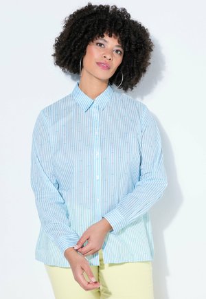 Kvinde med krøllet hår iført creoler, en lyseblå stribet skjorte med knapper og blege gule bukser, stående med hænderne foldet.