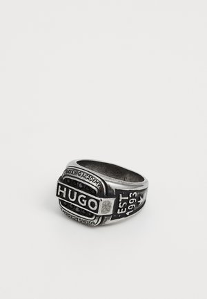 Anello in metallo color argento con parte superiore rettangolare che presenta le scritte "HUGO" e "BOXING ACADEMY" in rilievo, impreziosito da pietre nere. Dettaglio stabilito nel 1993.