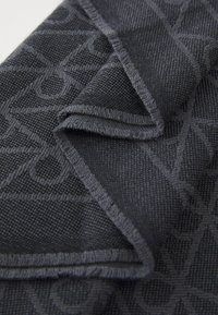 Grå tekstil med et geometrisk mønster og en tekstureret vævning. Stoffet har frynset kanter, hvilket skaber et lagdelt, foldet udseende.