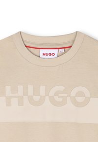 T-shirt en coton beige avec un col ras du cou, une texture lisse et un logo 'HUGO' en relief sur le devant dans une teinte contrastante.